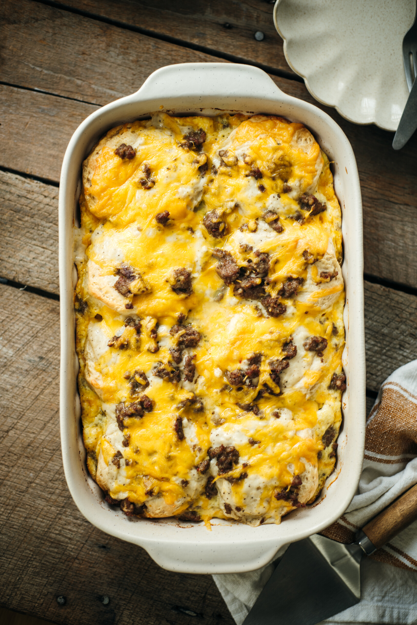Overhead view of a baked sausage biscuit casserole in a white dish, topped with melted cheddar cheese and browned sausage, resting on a rustic wooden table with a serving spatula nearby