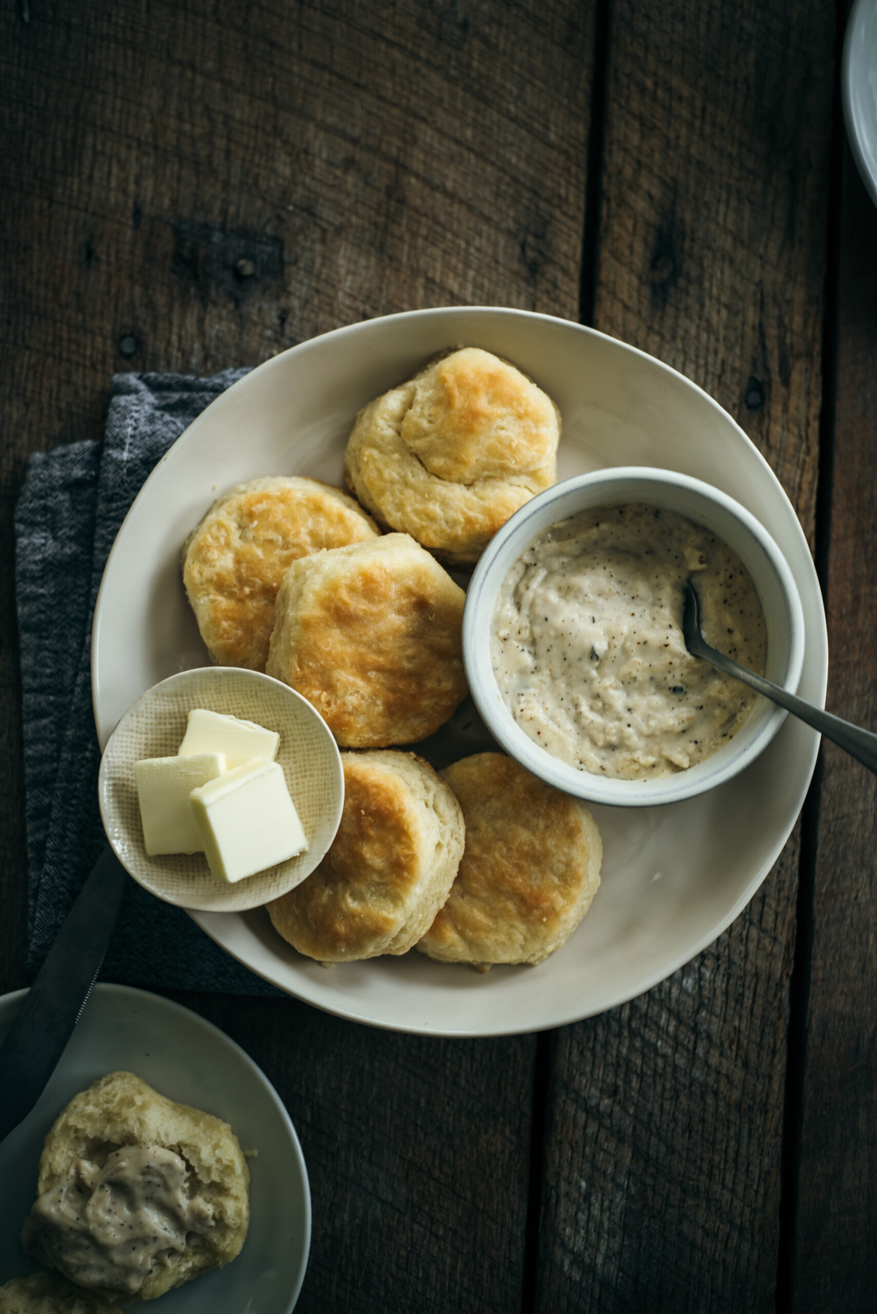Overhead view of biscuits and gravy served in a white bowl, accompanied by a small plate of butter and a biscuit topped with gravy.