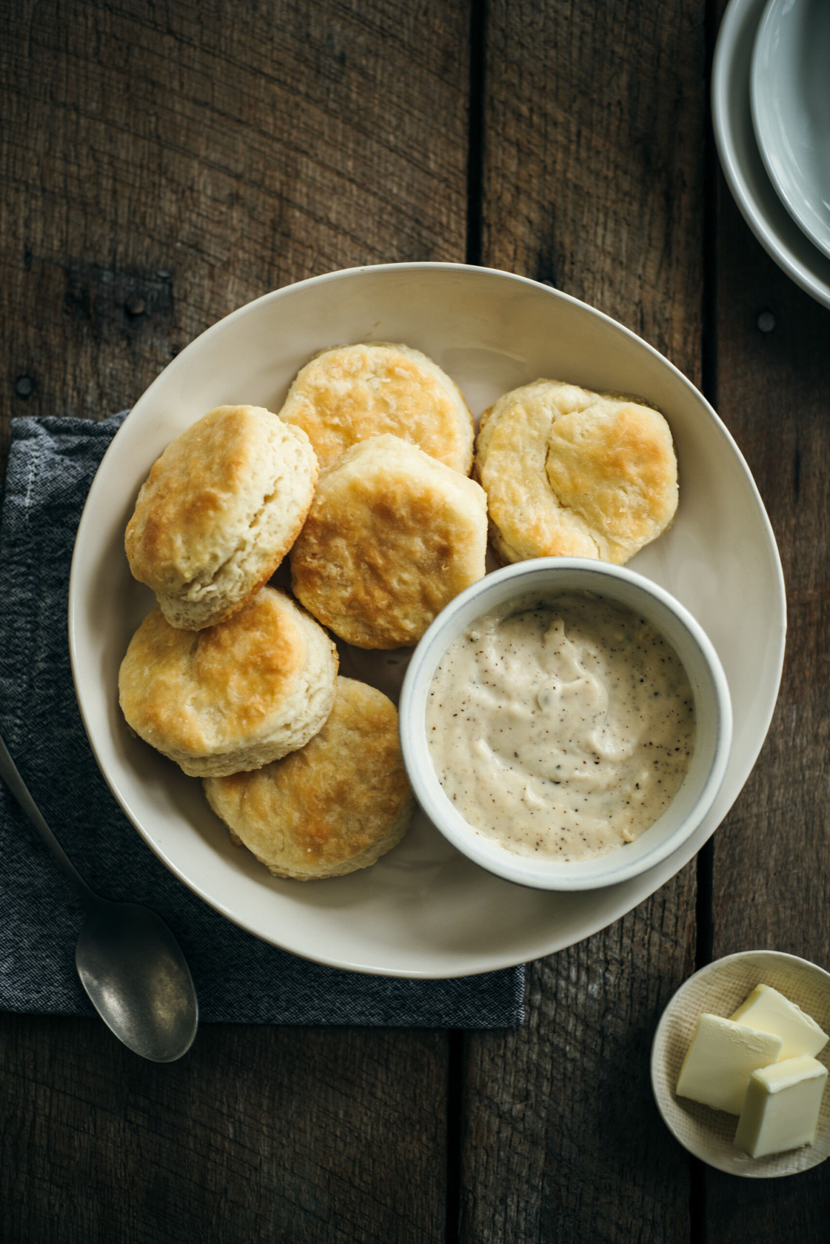 Overhead shot of a plate of homemade biscuits with a bowl of white pepper gravy, a spoon, and butter cubes on a rustic wooden surface.