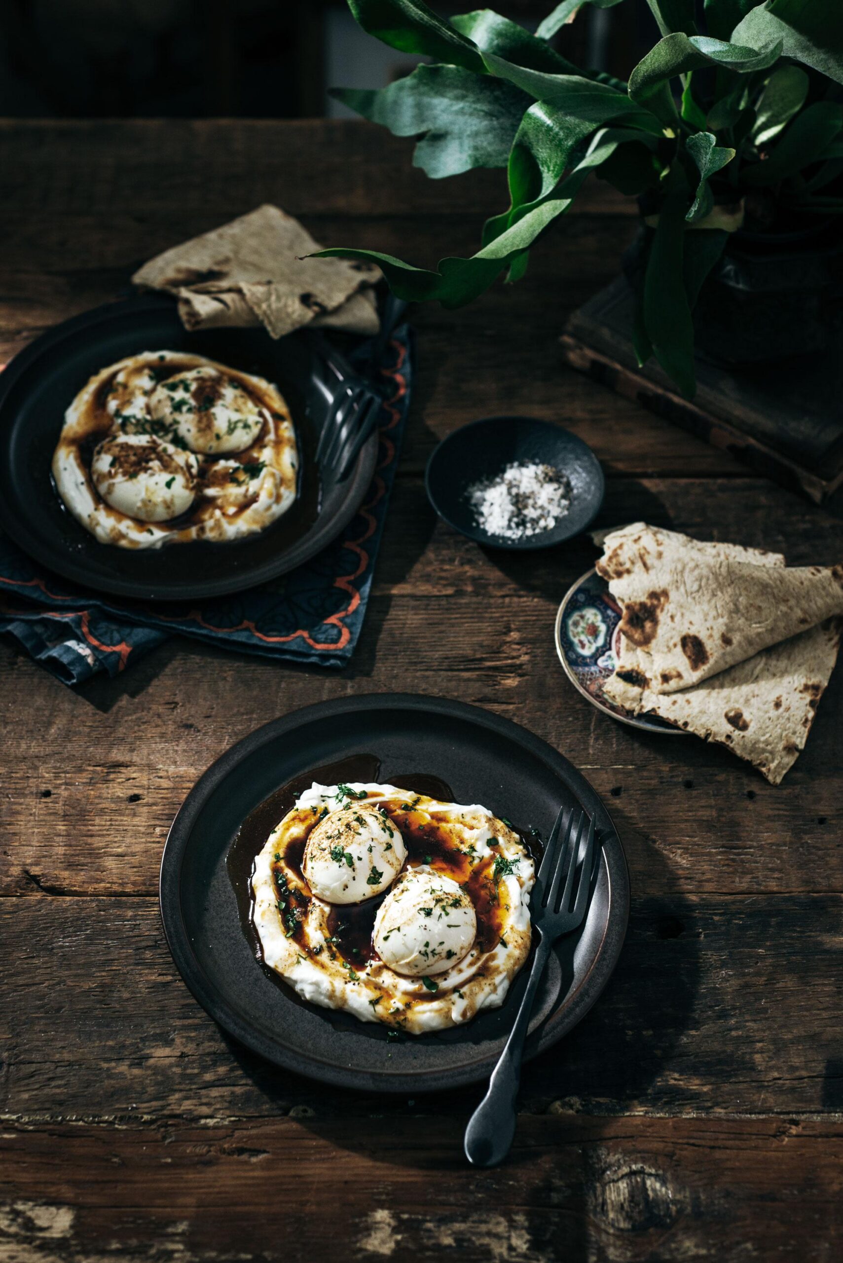 Two plates of poached eggs on creamy yogurt with herbs and sauce, served with flatbread on a rustic wooden table. A small bowl of salt and a green potted plant are also visible.