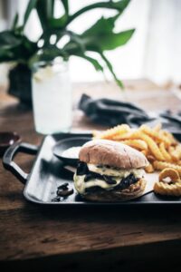 A cheeseburger with melted cheese and mushrooms sits on a tray next to crinkle-cut fries and onion rings, with a glass of water and leafy plant in the background on a wooden table.