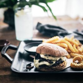 A cheeseburger with melted cheese and mushrooms sits on a tray next to crinkle-cut fries and onion rings, with a glass of water and leafy plant in the background on a wooden table.