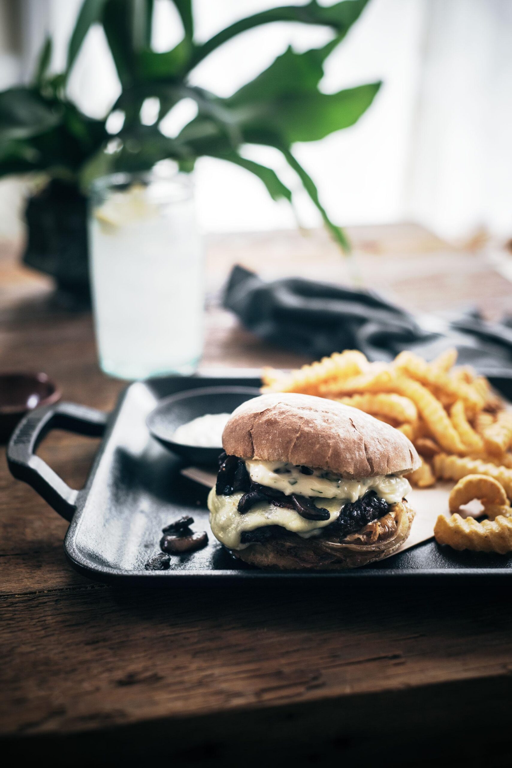 A cheeseburger with melted cheese and mushrooms sits on a tray next to crinkle-cut fries and onion rings, with a glass of water and leafy plant in the background on a wooden table.