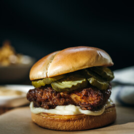 A portrait view of a honey butter chicken sandwich with fried chicken, pickles, and creamy sauce, placed on parchment paper with a blurred background