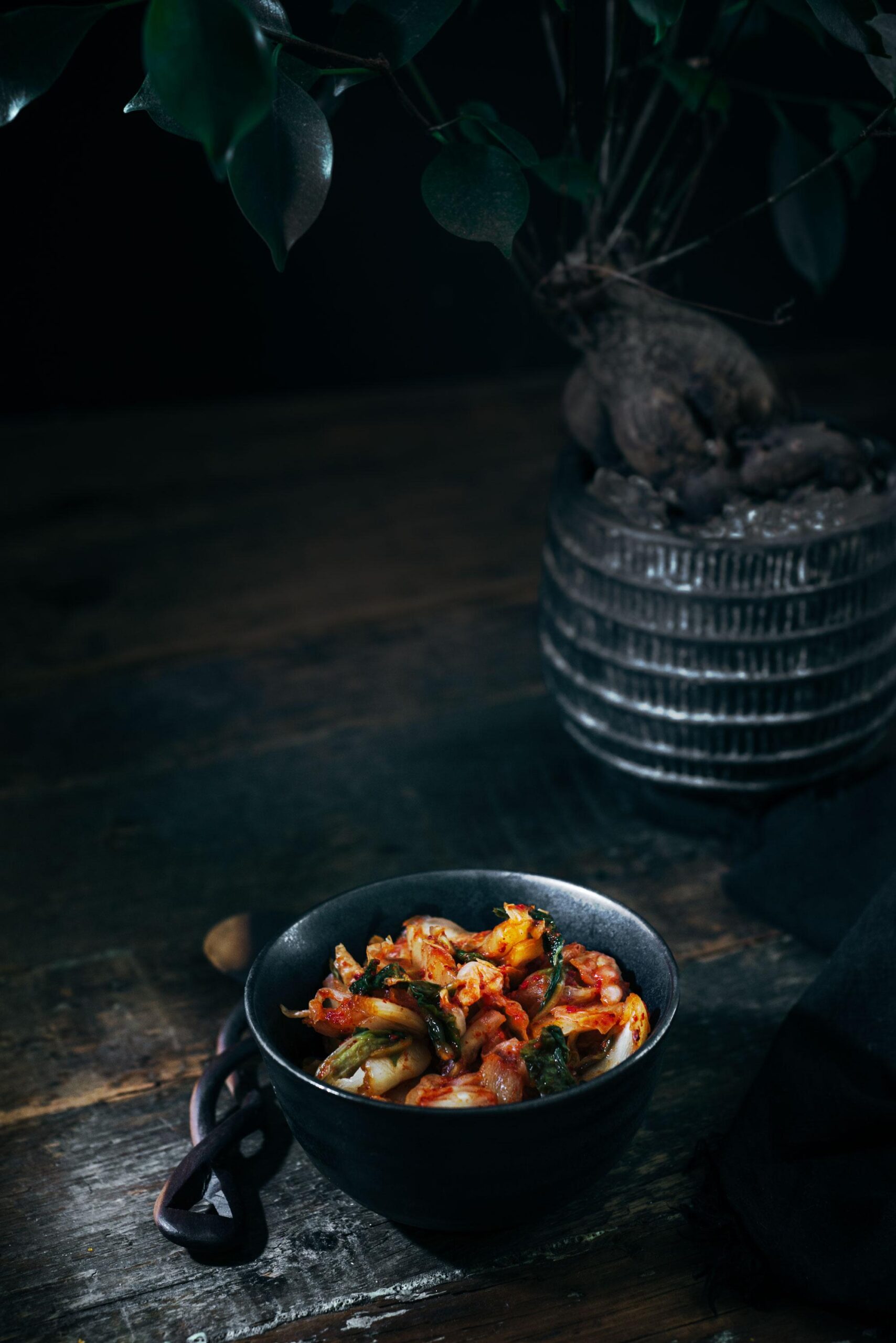 A dark bowl filled with kimchi sits on a rustic wooden table. In the background, a textured plant pot with a leafy green plant adds depth to the moody, dimly lit setting.