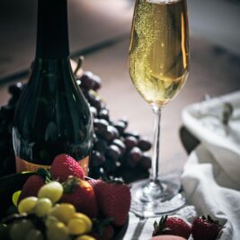 A glass of sparkling wine sits next to a dark wine bottle, with fresh strawberries, grapes, and a peach arranged on a table with a white cloth. Bubbles rise in the glass.