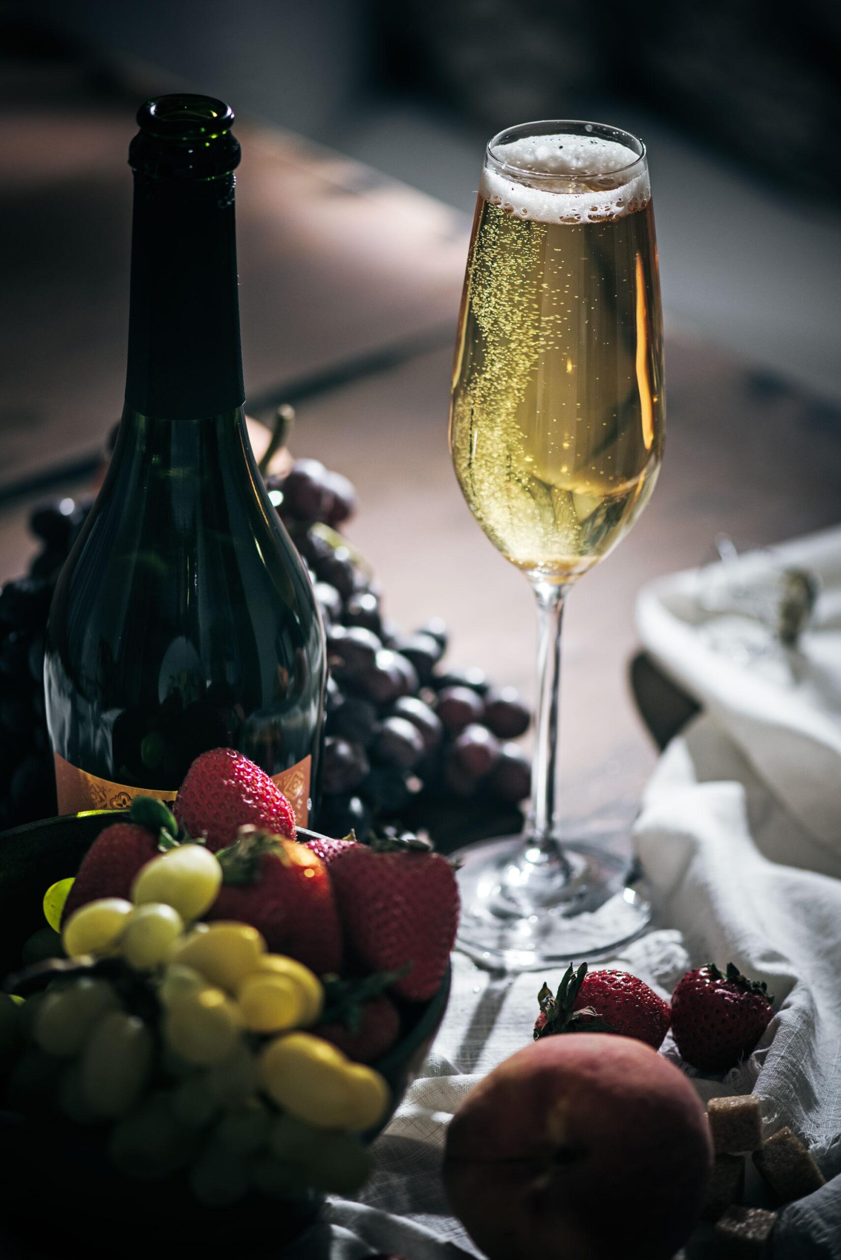A glass of sparkling wine sits next to a dark wine bottle, with fresh strawberries, grapes, and a peach arranged on a table with a white cloth. Bubbles rise in the glass.