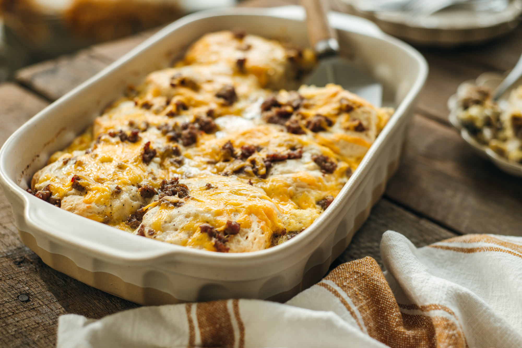 Freshly baked sausage biscuit casserole in a white baking dish, topped with melted cheese and browned sausage, with a serving spatula and a striped kitchen towel beside it.