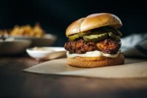 A horizontal shot of a honey butter chicken sandwich highlighting its golden bun, crispy chicken, pickles, and sauce, with side dishes blurred in the background on a wooden surface