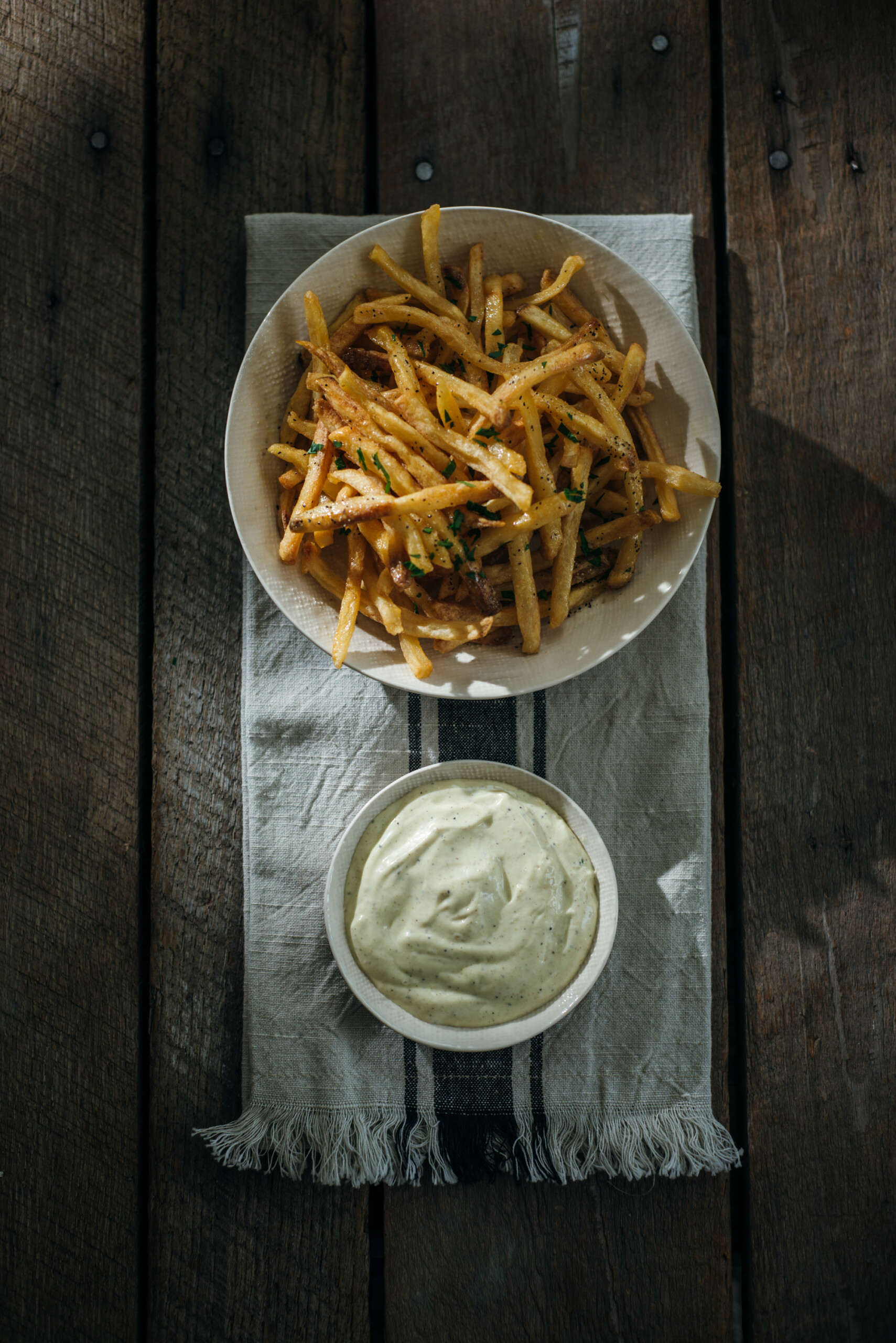 Overhead view of a bowl of thin, crispy fries garnished with herbs, served alongside a small bowl of creamy truffle aioli on a rustic wooden table with a striped cloth underneath.