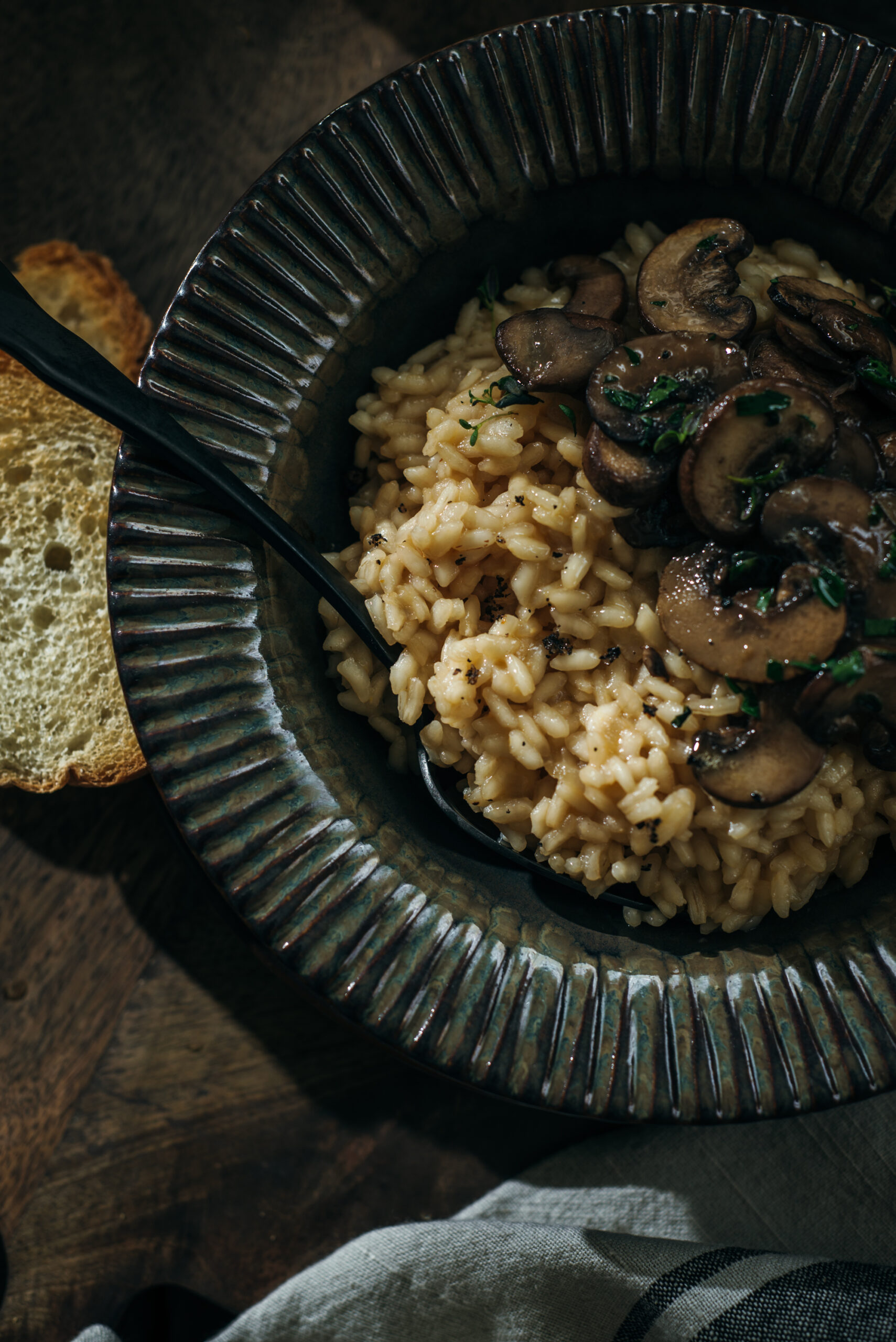 Close-up overhead view of creamy mushroom risotto topped with sautéed mushrooms and herbs, served in a textured bowl with a black spoon and a slice of toasted bread on the side