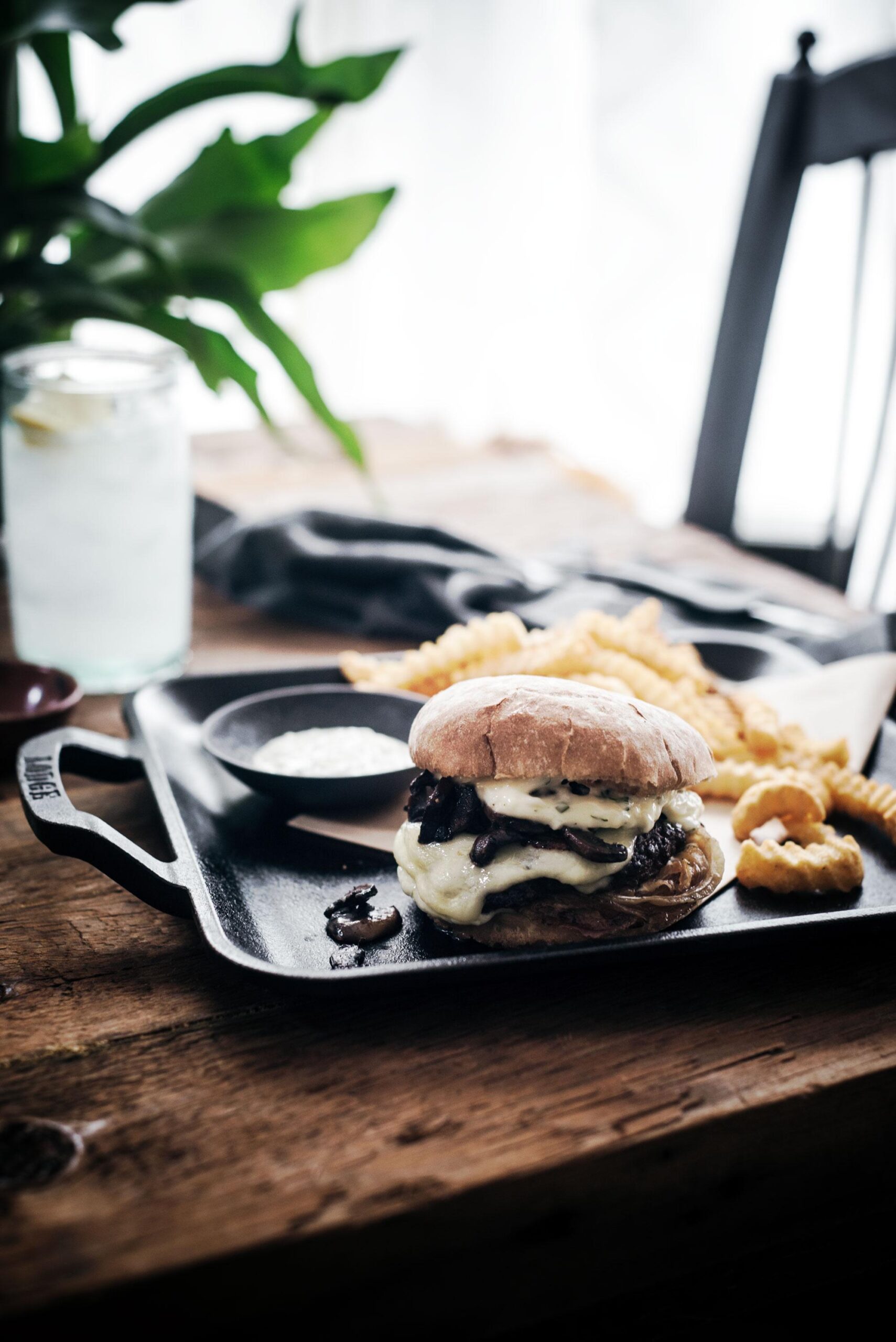 A cheeseburger with melted cheese and mushrooms sits on a black tray with crinkle-cut fries and dipping sauce, on a rustic wooden table. A glass of water with lemon and a leafy plant are in the background.