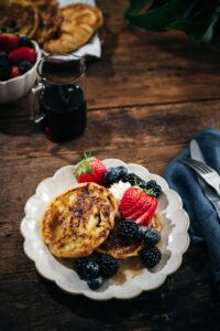 A plate with two pieces of French toast topped with whipped cream, strawberries, blackberries, and blueberries, sits on a wooden table beside a glass of dark syrup and a bowl of mixed berries.